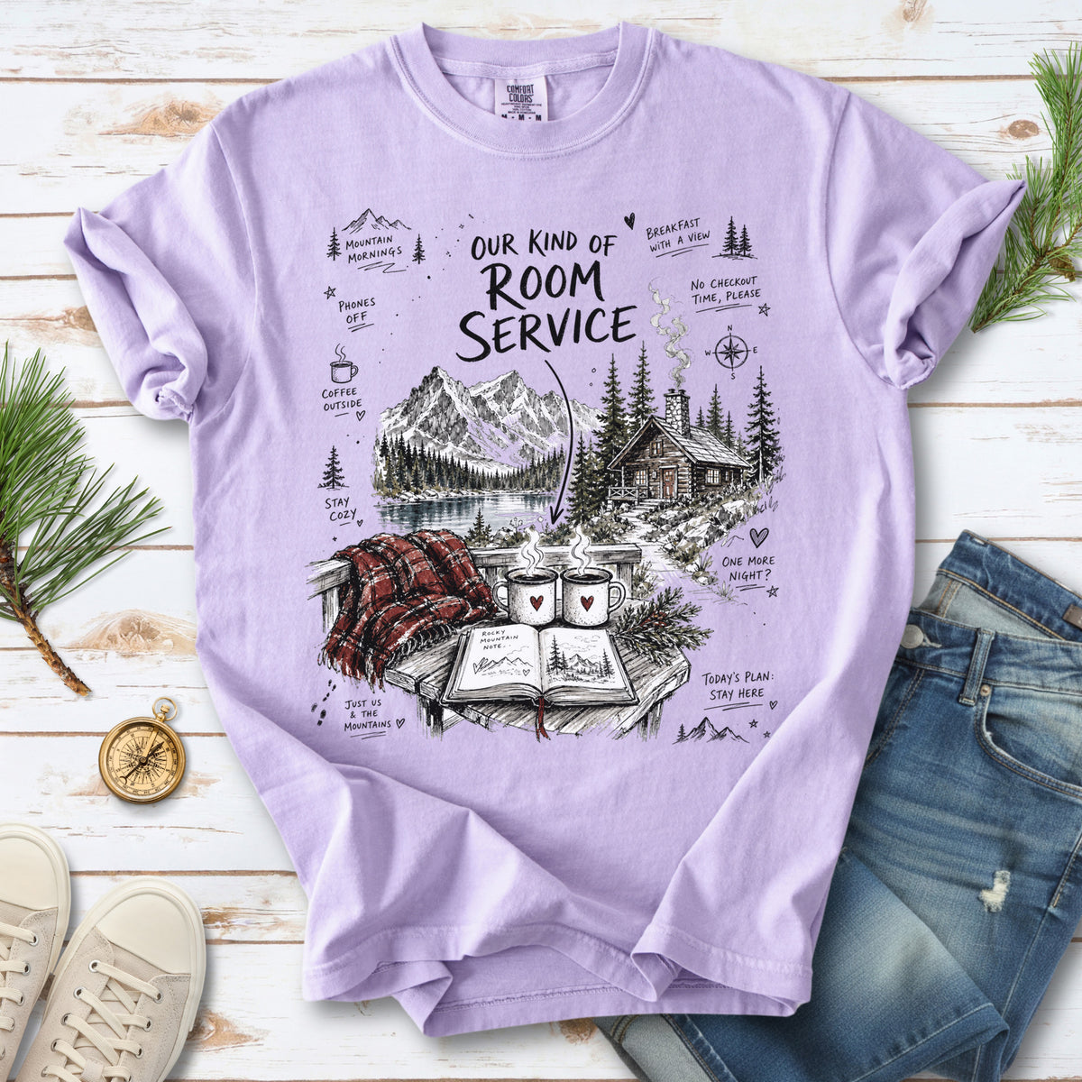 A cozy lakeside cabin scene with Rocky Mountain peaks, coffee mugs, and a journal design on an Orchid Comfort Colors T-shirt