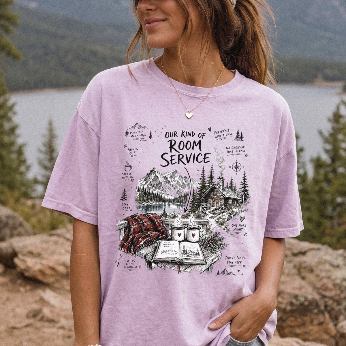 Woman wearing an Orchid Comfort Colors T-shirt printed with a cozy lakeside cabin scene with Rocky Mountain peaks, coffee mugs, and a journal