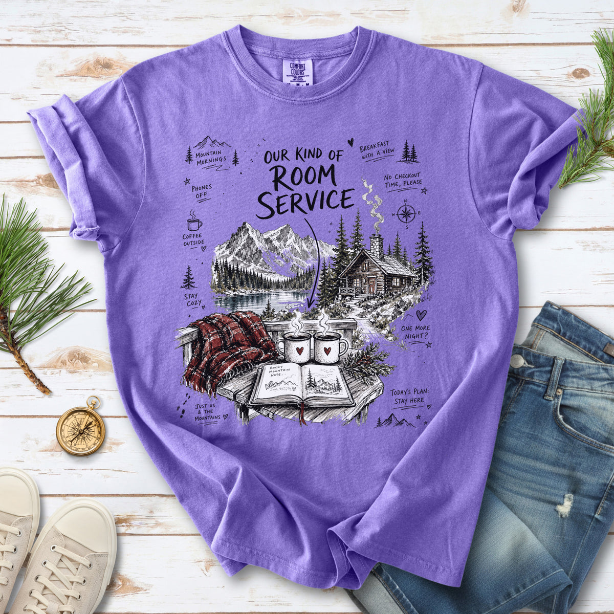 A cozy lakeside cabin scene with Rocky Mountain peaks, coffee mugs, and a journal design on a Violet Comfort Colors T-shirt