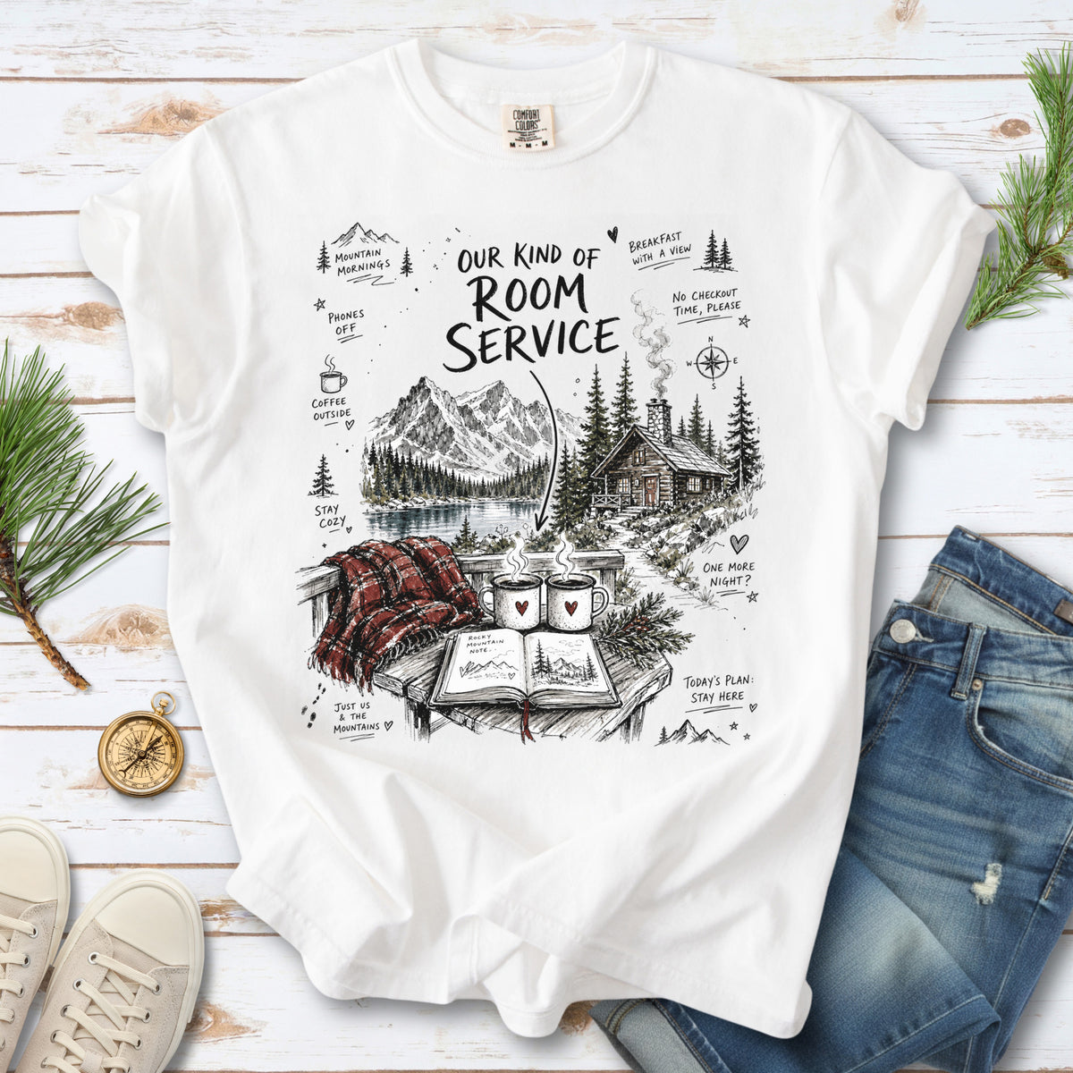 A cozy lakeside cabin scene with Rocky Mountain peaks, coffee mugs, and a journal design on a White Comfort Colors T-shirt