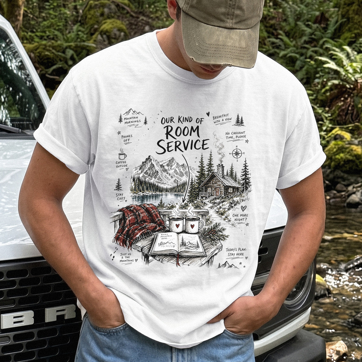 Man wearing a White Comfort Colors T-shirt with a cozy lakeside cabin scene with Rocky Mountain peaks, coffee mugs, and a journal