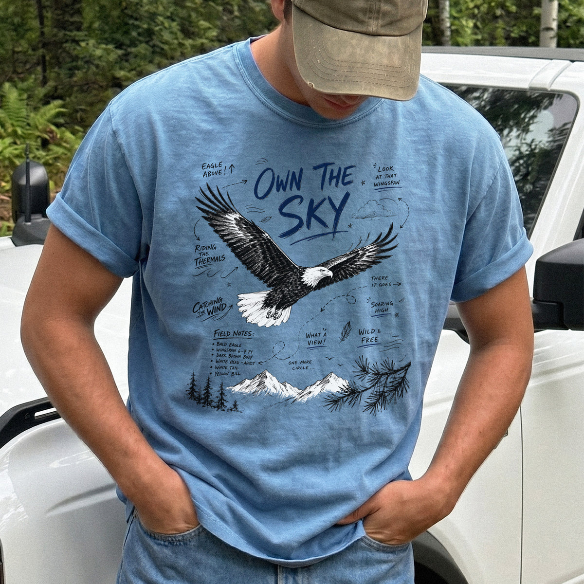 Own the Sky Comfort Colors Shirt – Bald Eagle design of a soaring Bald Eagle in flight above mountain peaks, with field-note style details highlighting its dark brown body, white head and tail, and yellow bill, printed on a Blue Jean T-shirt.