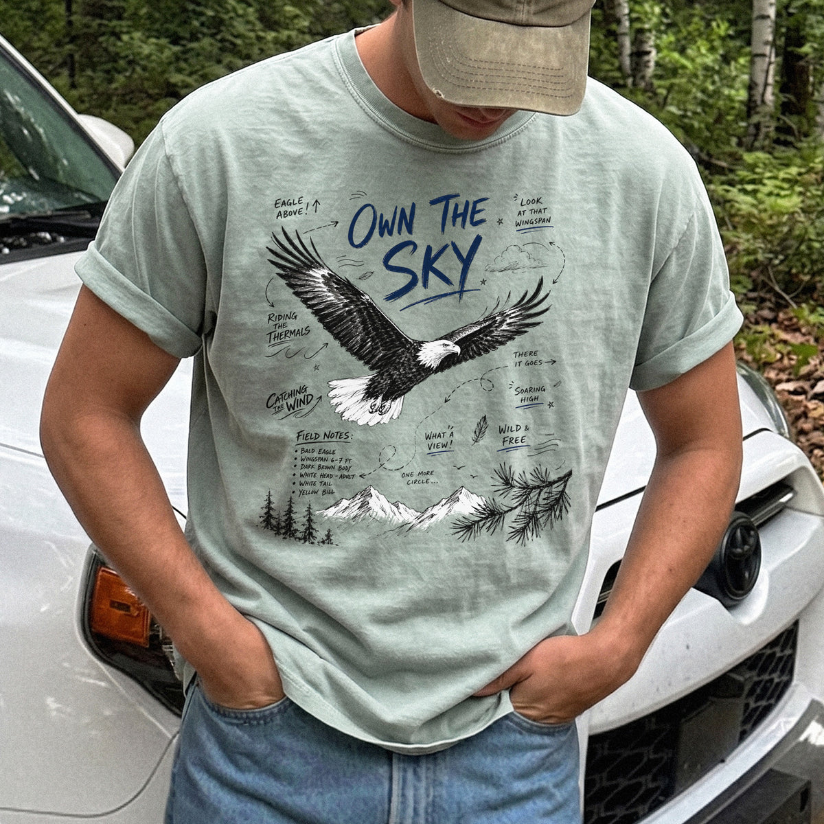 Own the Sky Comfort Colors Shirt – Bald Eagle design of a soaring Bald Eagle in flight above mountain peaks, with field-note style details highlighting its dark brown body, white head and tail, and yellow bill, printed on a Bay T-shirt.