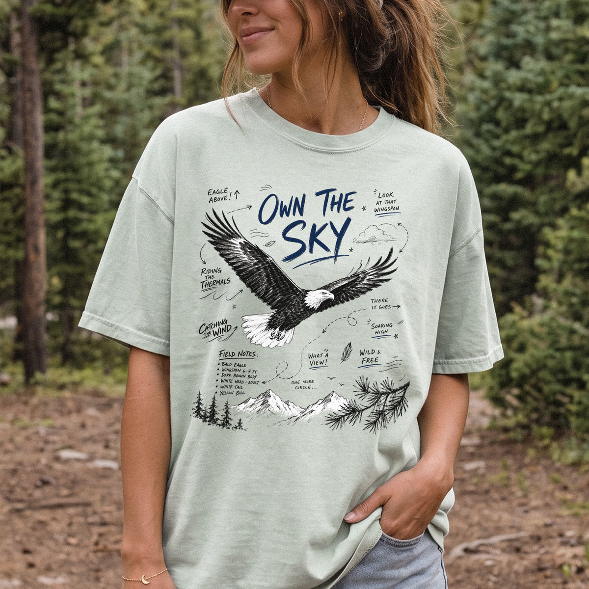 Own the Sky Comfort Colors Shirt – Bald Eagle design of a soaring Bald Eagle in flight above mountain peaks, with field-note style details highlighting its dark brown body, white head and tail, and yellow bill, printed on a Bay T-shirt, worn by a woman.