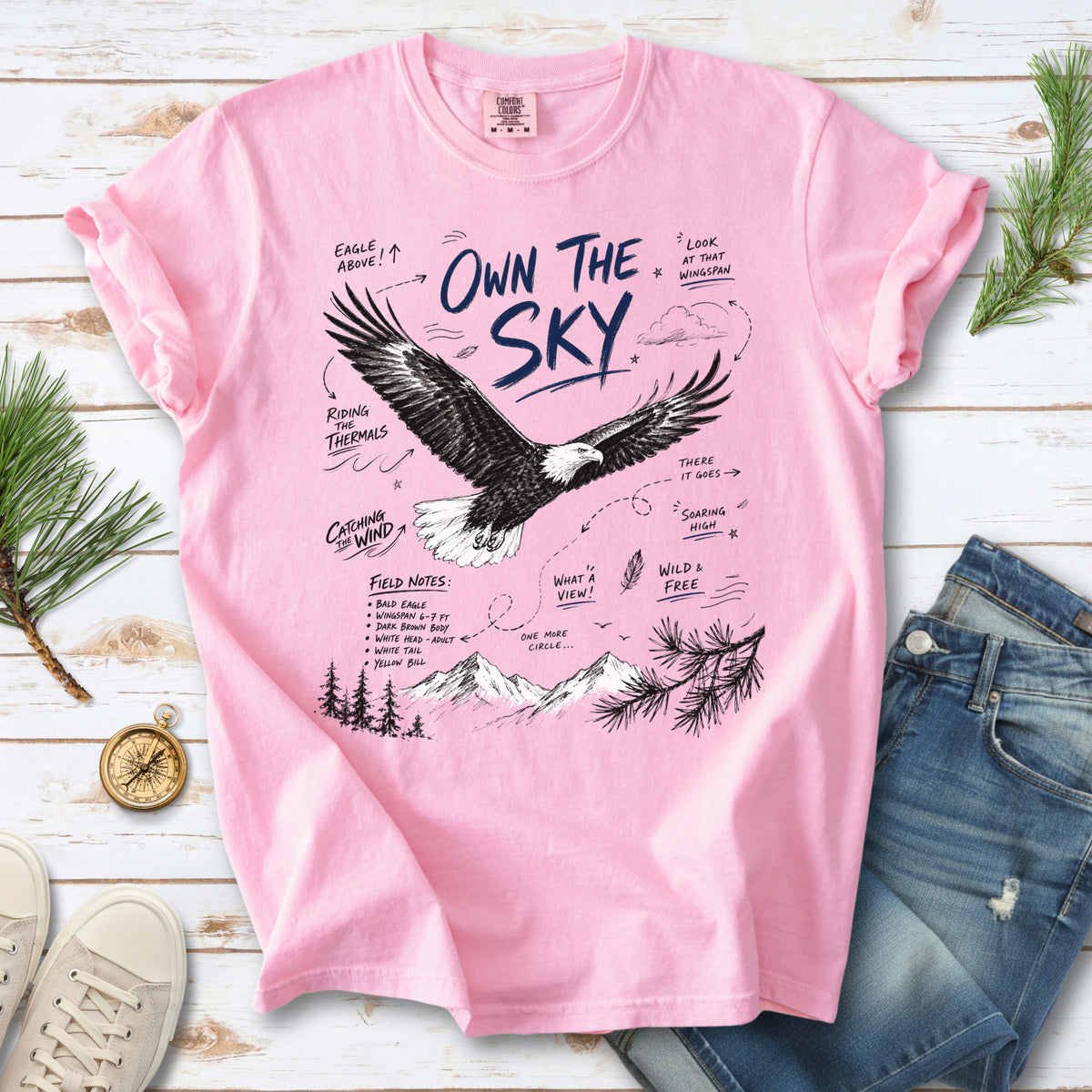 Own the Sky Comfort Colors Shirt – Bald Eagle design of a soaring Bald Eagle in flight above mountain peaks, with field-note style details highlighting its dark brown body, white head and tail, and yellow bill, printed on a Blossom T-shirt.