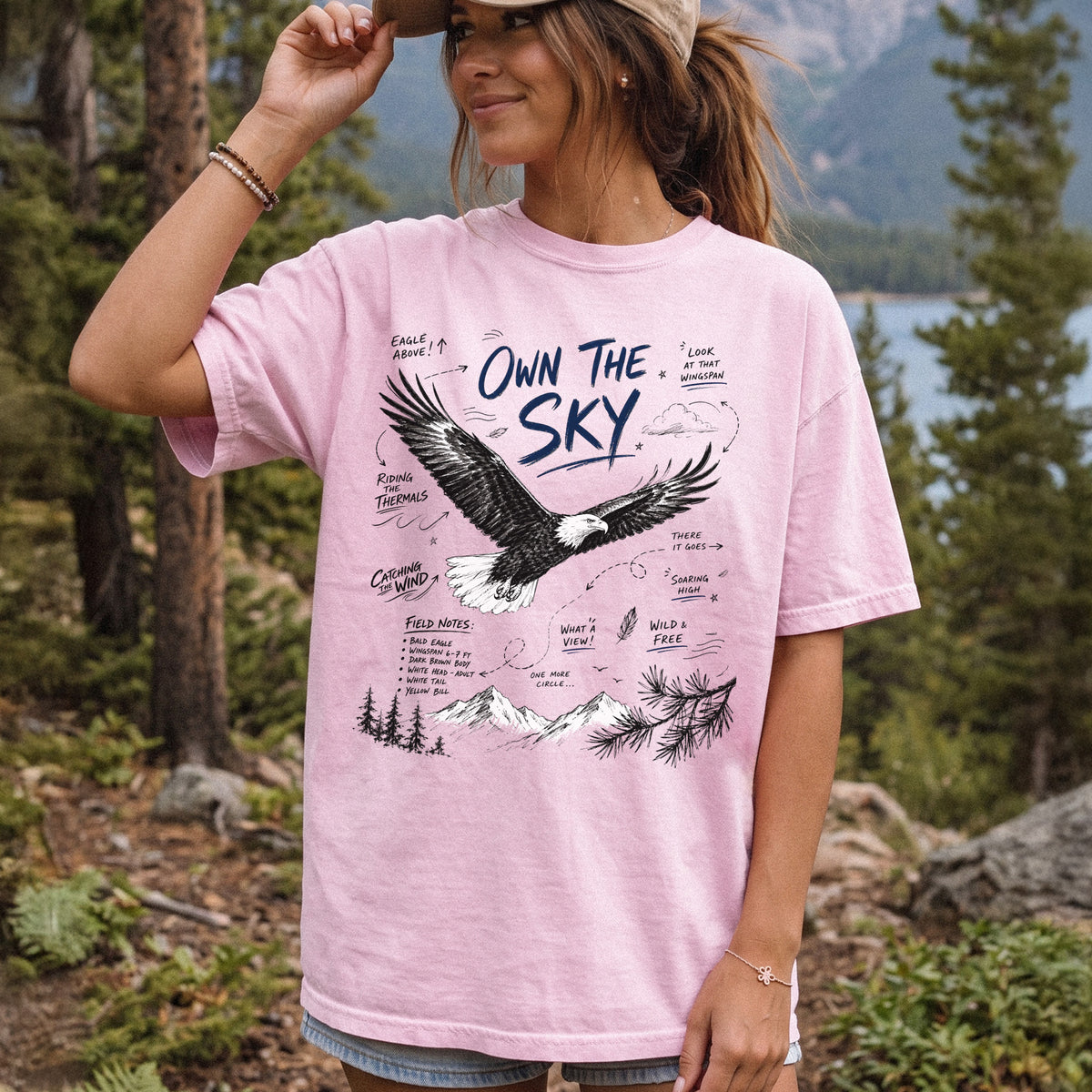 Own the Sky Comfort Colors Shirt – Bald Eagle design of a soaring Bald Eagle in flight above mountain peaks, with field-note style details highlighting its dark brown body, white head and tail, and yellow bill, printed on a Blossom T-shirt, worn by a woman.