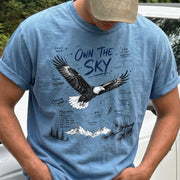 Own the Sky Comfort Colors Shirt – Bald Eagle design of a soaring Bald Eagle in flight above mountain peaks, with field-note style details highlighting its dark brown body, white head and tail, and yellow bill, printed on a Blue Jean T-shirt.