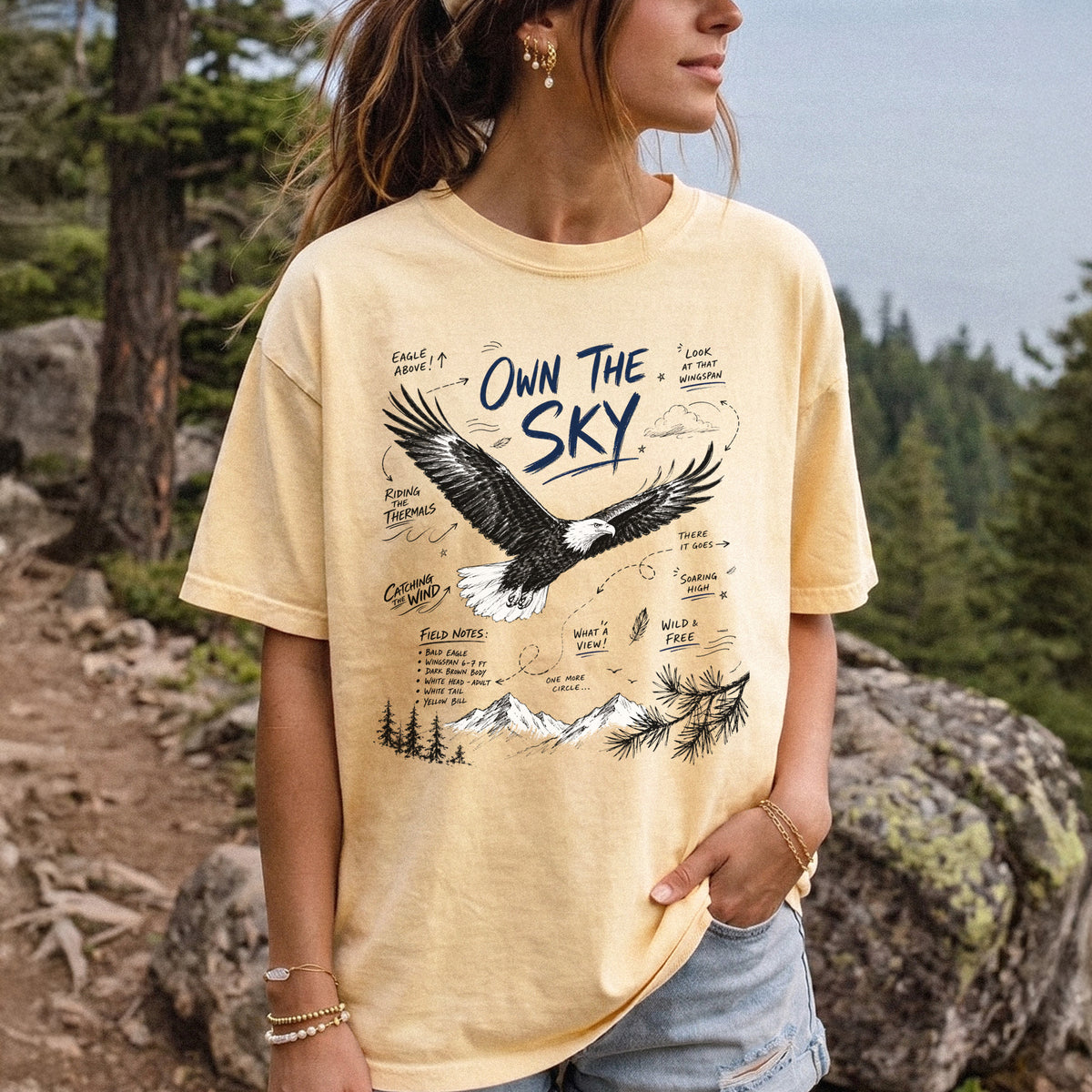 Own the Sky Comfort Colors Shirt – Bald Eagle design of a soaring Bald Eagle in flight above mountain peaks, with field-note style details highlighting its dark brown body, white head and tail, and yellow bill, printed on a Butter T-shirt, worn by a woman.