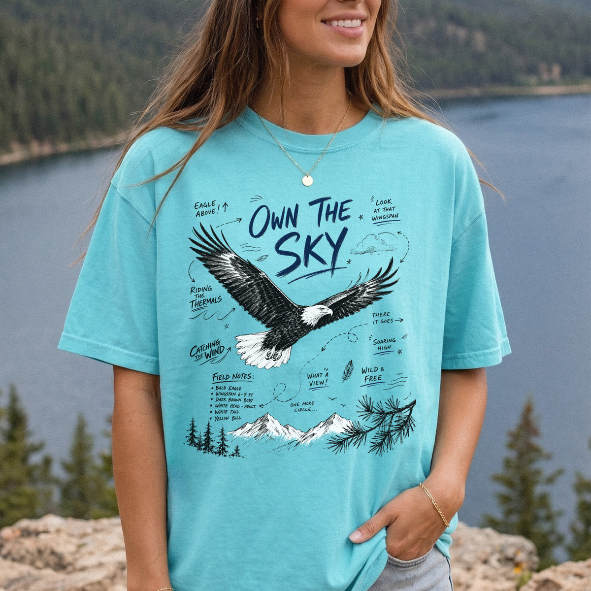 Own the Sky Comfort Colors Shirt – Bald Eagle design of a soaring Bald Eagle in flight above mountain peaks, with field-note style details highlighting its dark brown body, white head and tail, and yellow bill, printed on a Chalky Mint T-shirt, worn by a woman.