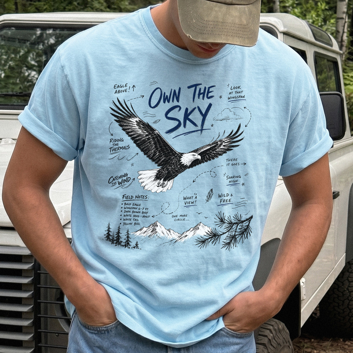 Own the Sky Comfort Colors Shirt – Bald Eagle design of a soaring Bald Eagle in flight above mountain peaks, with field-note style details highlighting its dark brown body, white head and tail, and yellow bill, printed on a Chambray T-shirt.