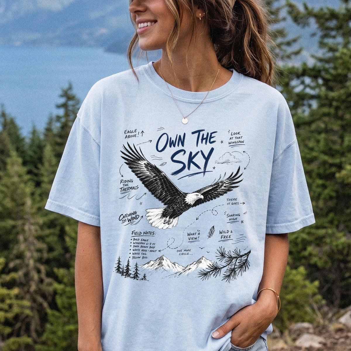 Own the Sky Comfort Colors Shirt – Bald Eagle design of a soaring Bald Eagle in flight above mountain peaks, with field-note style details highlighting its dark brown body, white head and tail, and yellow bill, printed on a Chambray T-shirt, worn by a woman.