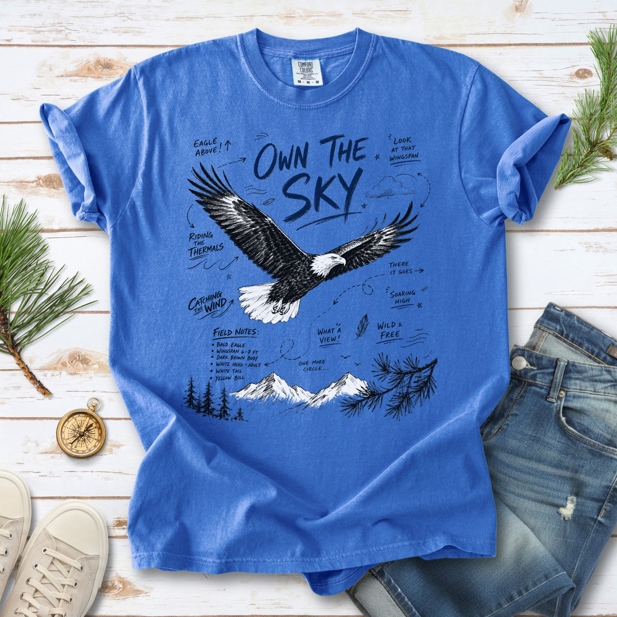 Own the Sky Comfort Colors Shirt – Bald Eagle design of a soaring Bald Eagle in flight above mountain peaks, with field-note style details highlighting its dark brown body, white head and tail, and yellow bill, printed on a Flo Blue T-shirt.