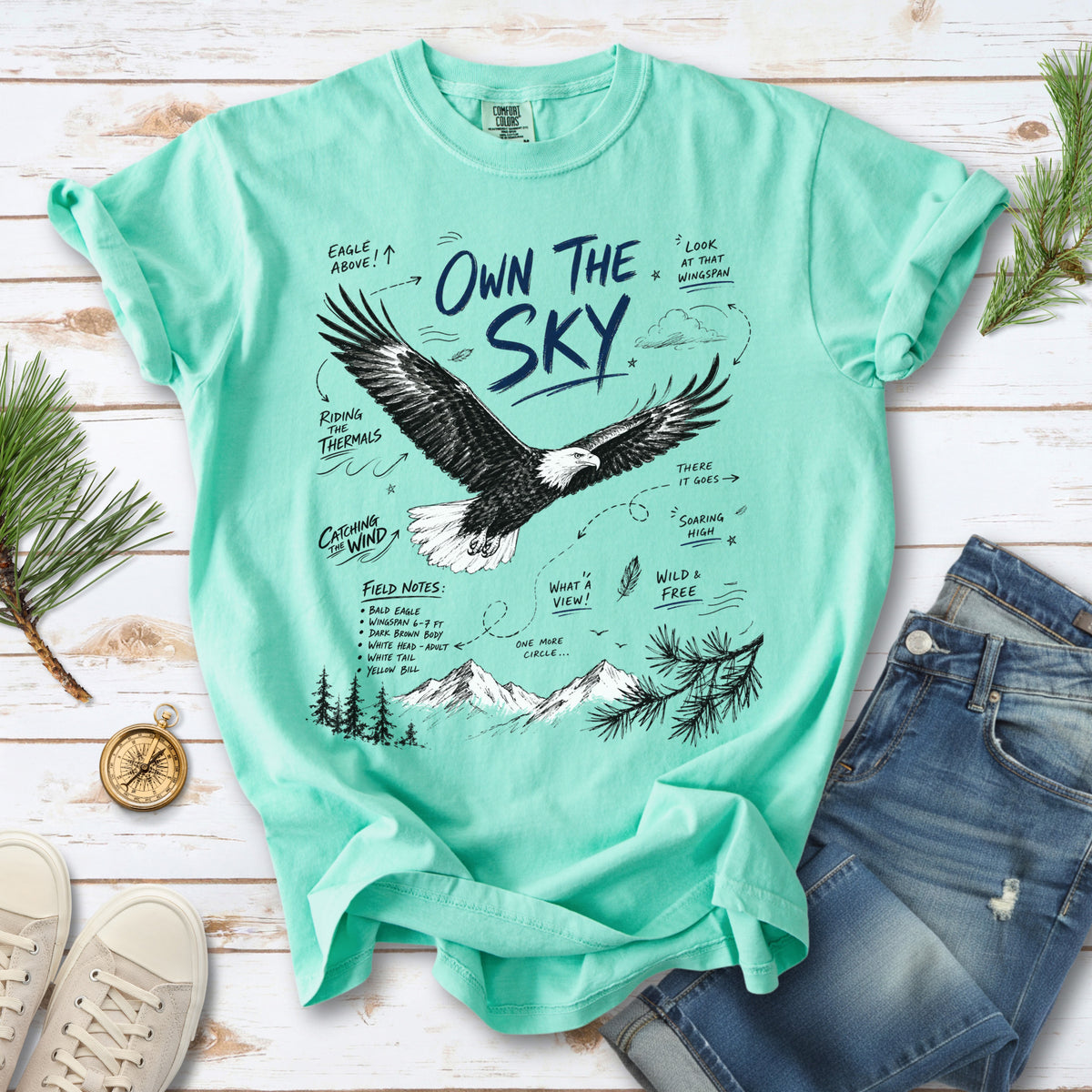 Own the Sky Comfort Colors Shirt – Bald Eagle design of a soaring Bald Eagle in flight above mountain peaks, with field-note style details highlighting its dark brown body, white head and tail, and yellow bill, printed on an Island Reef T-shirt.