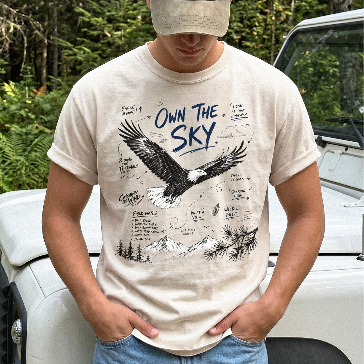 Own the Sky Comfort Colors Shirt – Bald Eagle design of a soaring Bald Eagle in flight above mountain peaks, with field-note style details highlighting its dark brown body, white head and tail, and yellow bill, printed on an Ivory T-shirt.