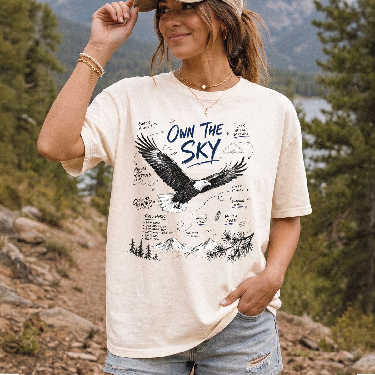 Own the Sky Comfort Colors Shirt – Bald Eagle design of a soaring Bald Eagle in flight above mountain peaks, with field-note style details highlighting its dark brown body, white head and tail, and yellow bill, printed on an Ivory T-shirt, worn by a woman.