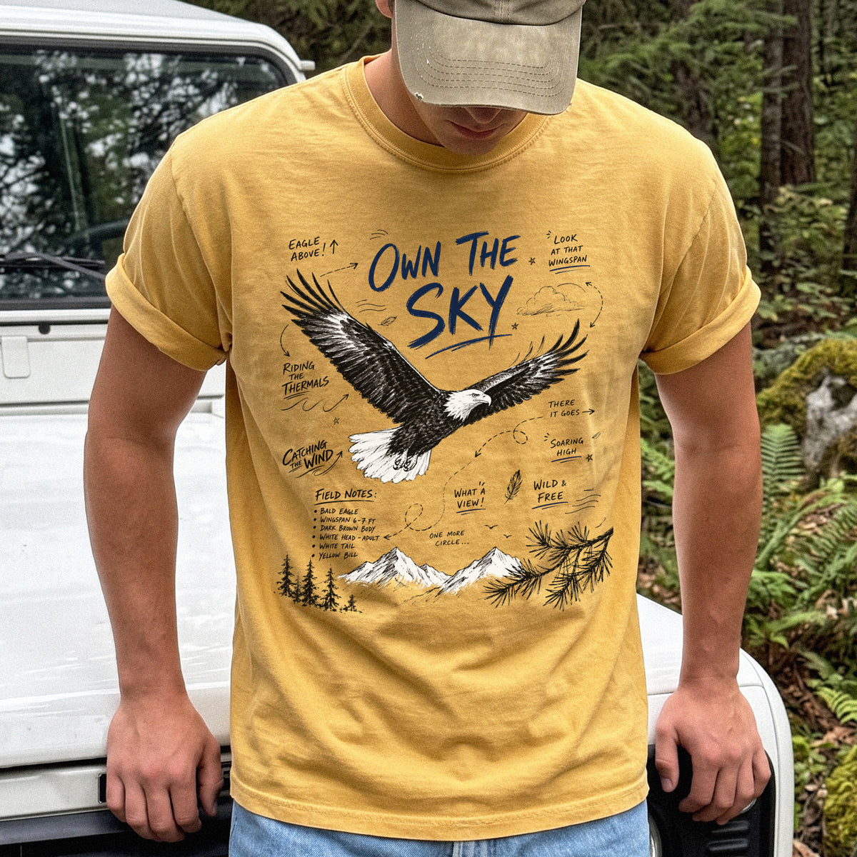Own the Sky Comfort Colors Shirt – Bald Eagle design of a soaring Bald Eagle in flight above mountain peaks, with field-note style details highlighting its dark brown body, white head and tail, and yellow bill, printed on a Mustard T-shirt.