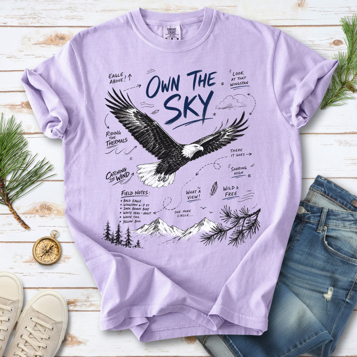 Own the Sky Comfort Colors Shirt – Bald Eagle design of a soaring Bald Eagle in flight above mountain peaks, with field-note style details highlighting its dark brown body, white head and tail, and yellow bill, printed on an Orchid T-shirt.