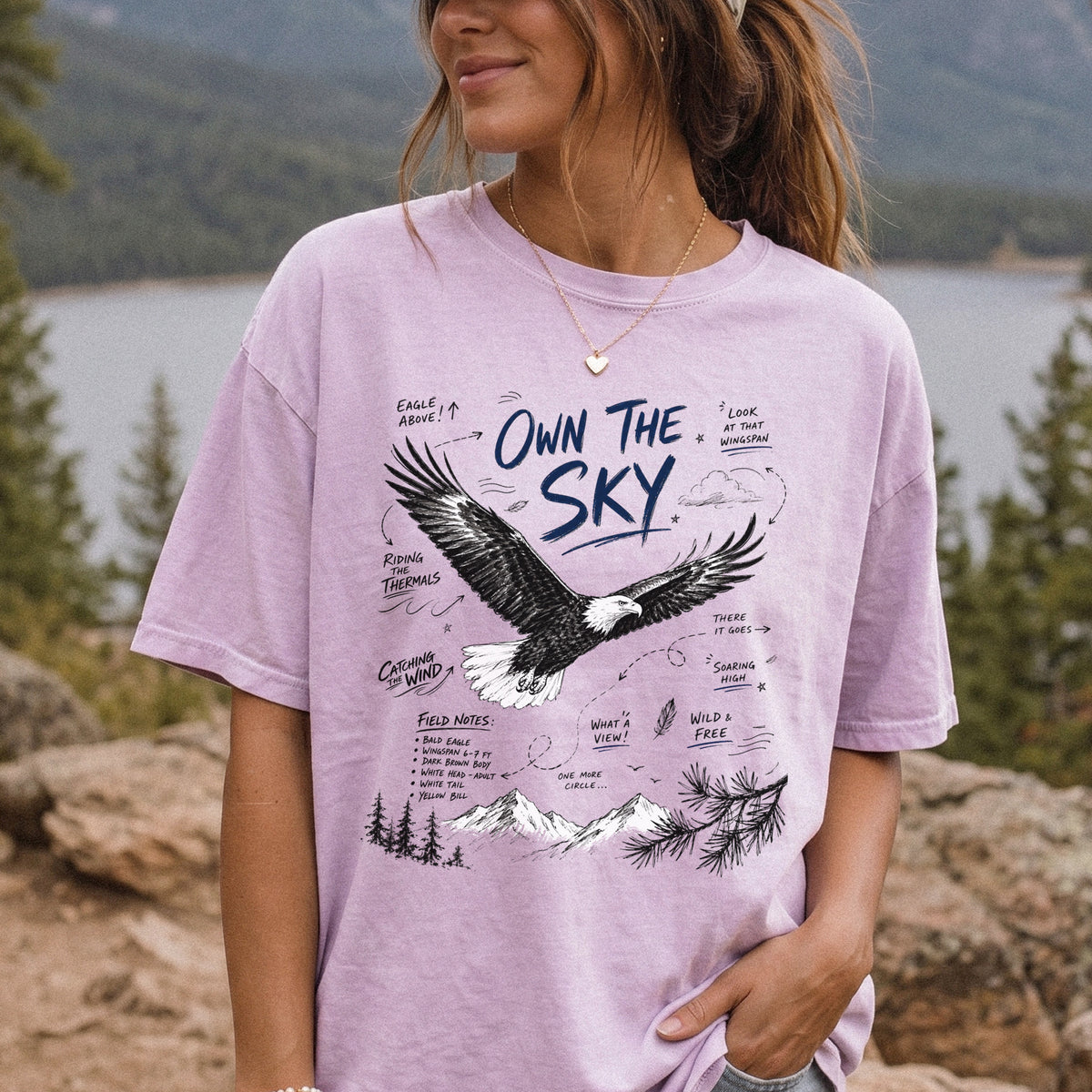 Own the Sky Comfort Colors Shirt – Bald Eagle design of a soaring Bald Eagle in flight above mountain peaks, with field-note style details highlighting its dark brown body, white head and tail, and yellow bill, printed on an Orchid T-shirt, worn by a woman.
