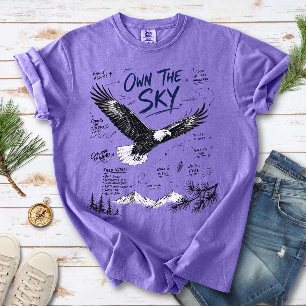 Own the Sky Comfort Colors Shirt – Bald Eagle design of a soaring Bald Eagle in flight above mountain peaks, with field-note style details highlighting its dark brown body, white head and tail, and yellow bill, printed on a Violet T-shirt.