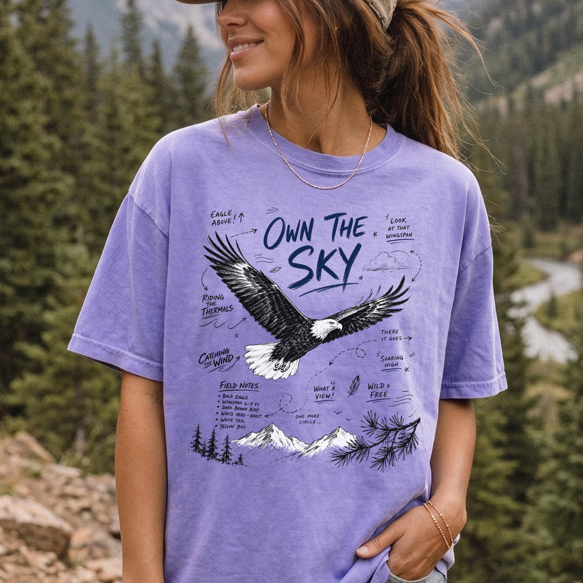 Own the Sky Comfort Colors Shirt – Bald Eagle design of a soaring Bald Eagle in flight above mountain peaks, with field-note style details highlighting its dark brown body, white head and tail, and yellow bill, printed on a Violet T-shirt, worn by a woman.