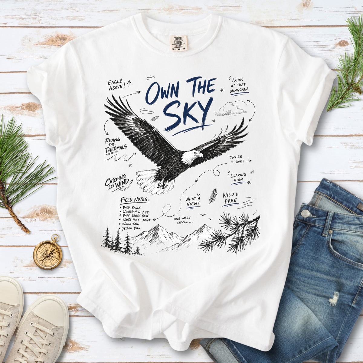 Own the Sky Comfort Colors Shirt – Bald Eagle design of a soaring Bald Eagle in flight above mountain peaks, with field-note style details highlighting its dark brown body, white head and tail, and yellow bill, printed on a White T-shirt.