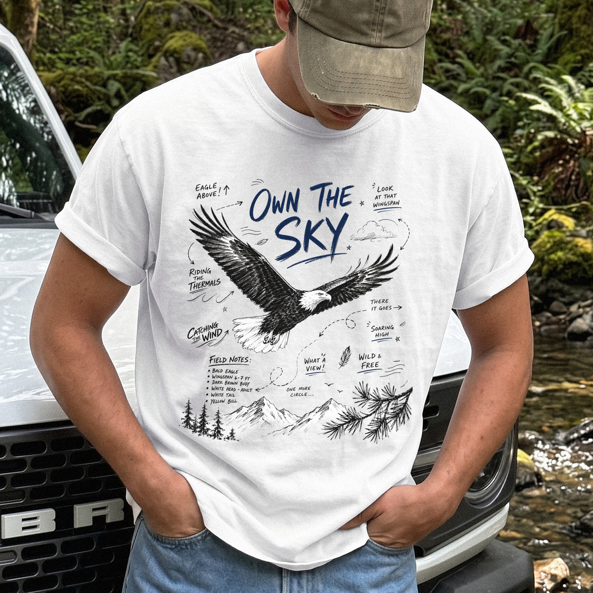 Own the Sky Comfort Colors Shirt – Bald Eagle design of a soaring Bald Eagle in flight above mountain peaks, with field-note style details highlighting its dark brown body, white head and tail, and yellow bill, printed on a White T-shirt.