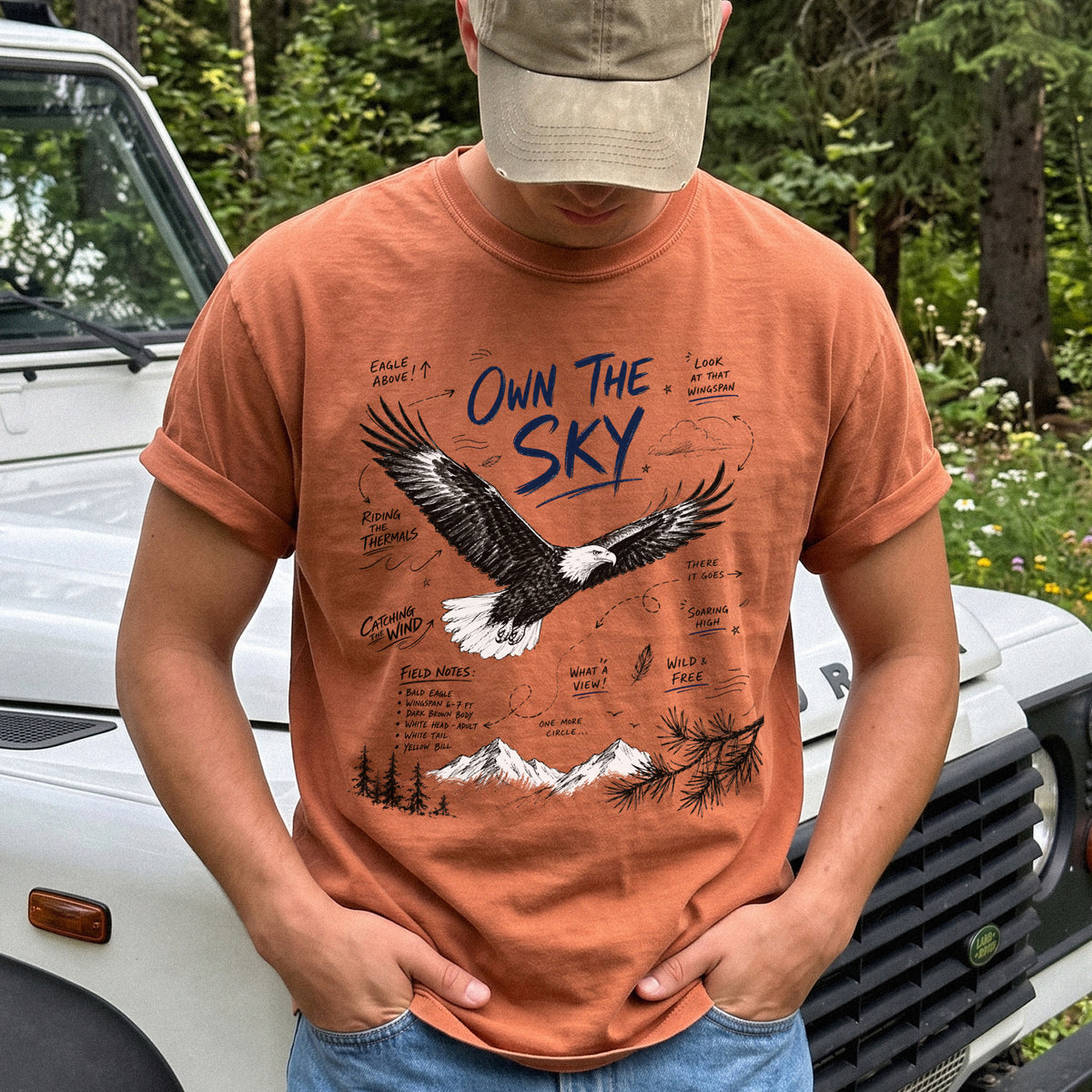 Own the Sky Comfort Colors Shirt – Bald Eagle design of a soaring Bald Eagle in flight above mountain peaks, with field-note style details highlighting its dark brown body, white head and tail, and yellow bill, printed on a Yam T-shirt.