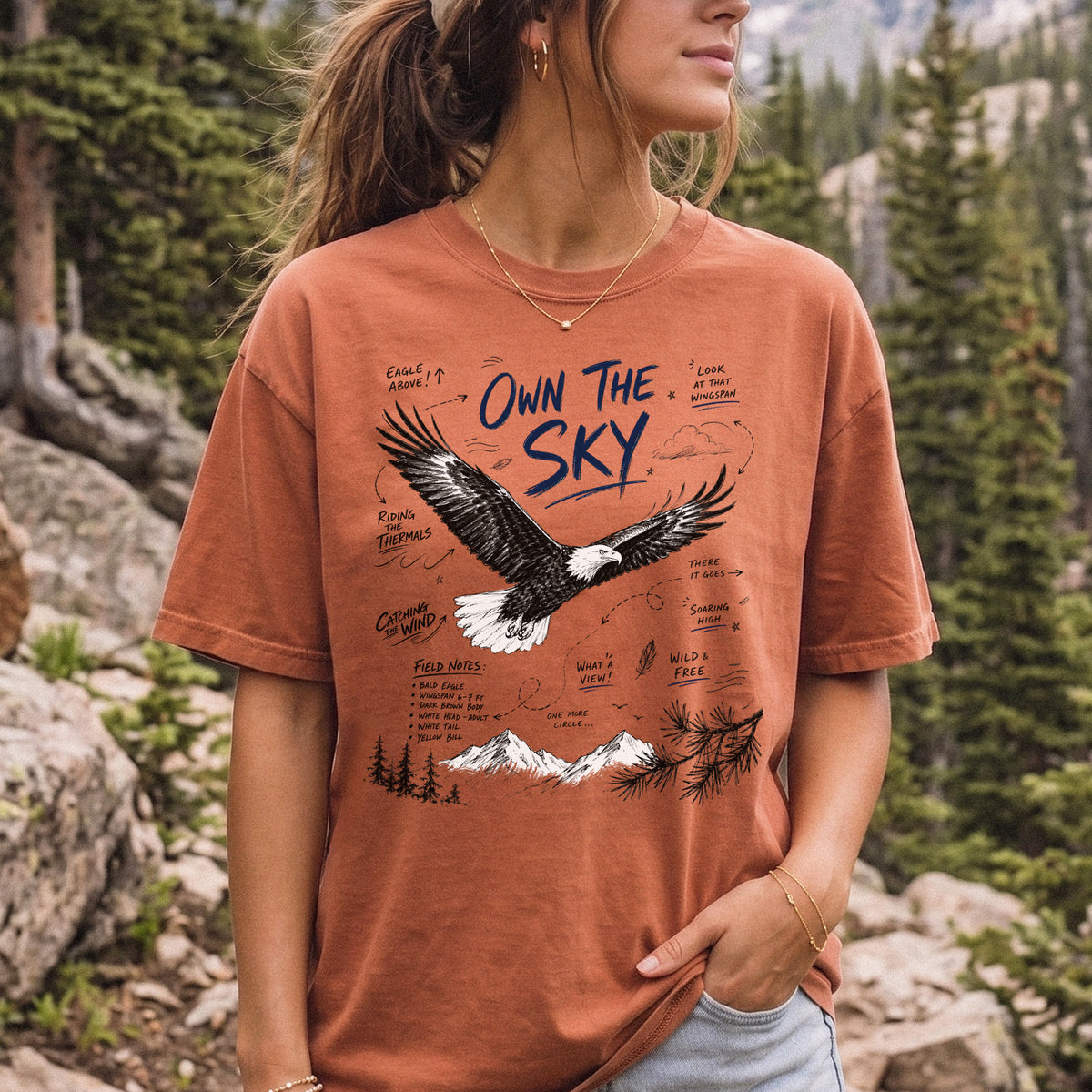 Own the Sky Comfort Colors Shirt – Bald Eagle design of a soaring Bald Eagle in flight above mountain peaks, with field-note style details highlighting its dark brown body, white head and tail, and yellow bill, printed on a Yam T-shirt, worn by a woman.