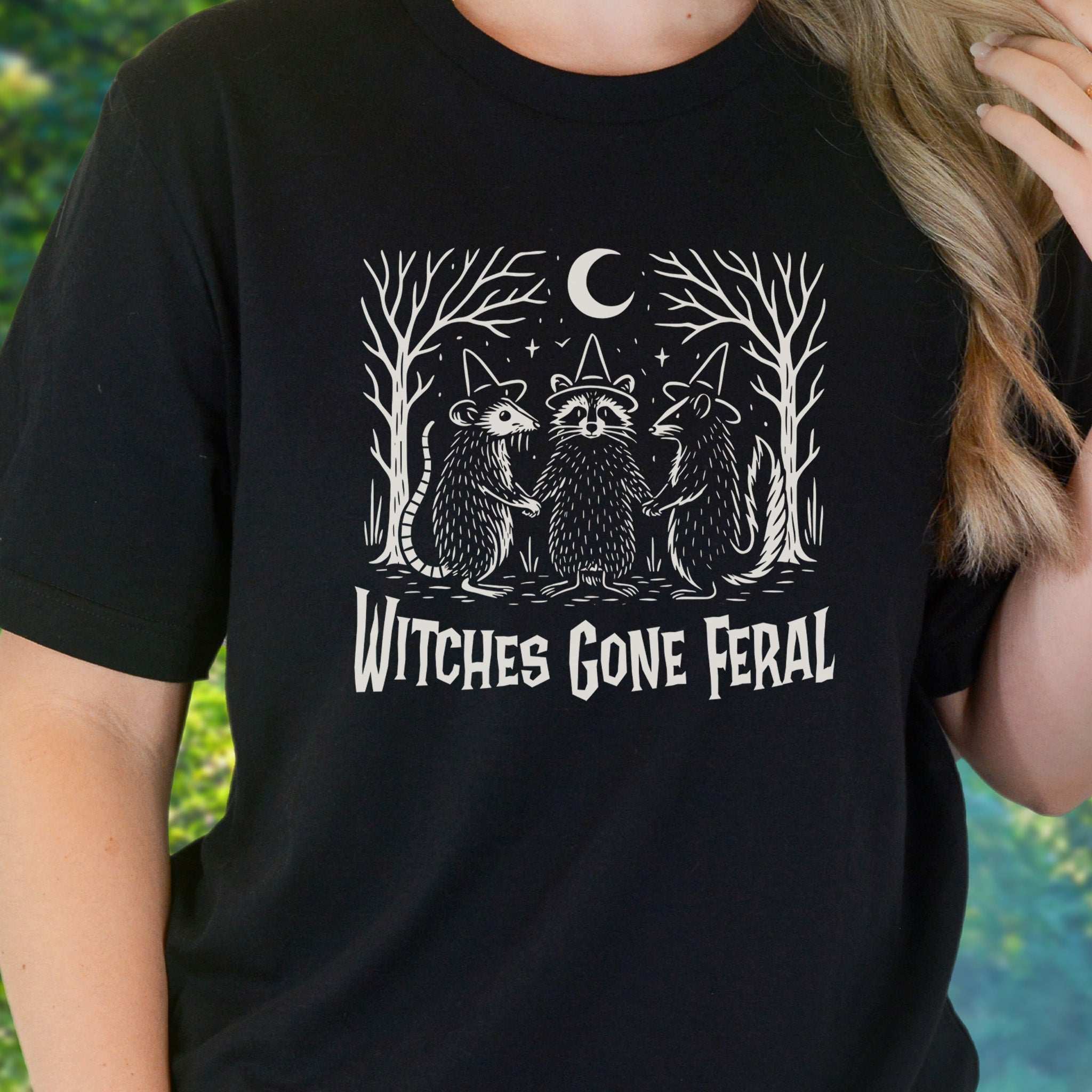 Witches Gone Feral Halloween T-shirt featuring raccoon, opossum, and skunk in a mystical forest scene.