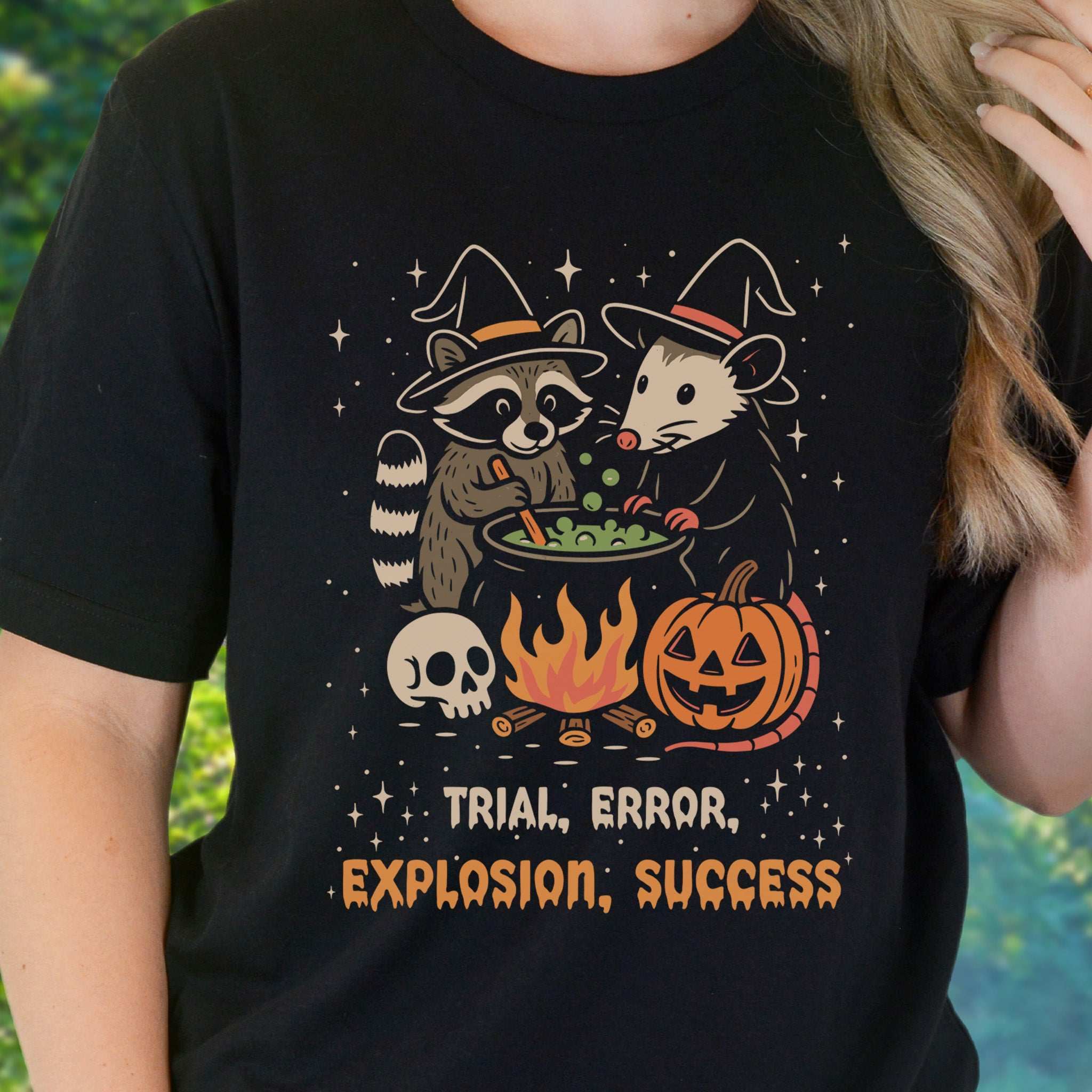 Potion Explosion Halloween T-shirt featuring raccoon and opossum witches in a cauldron with pumpkins.