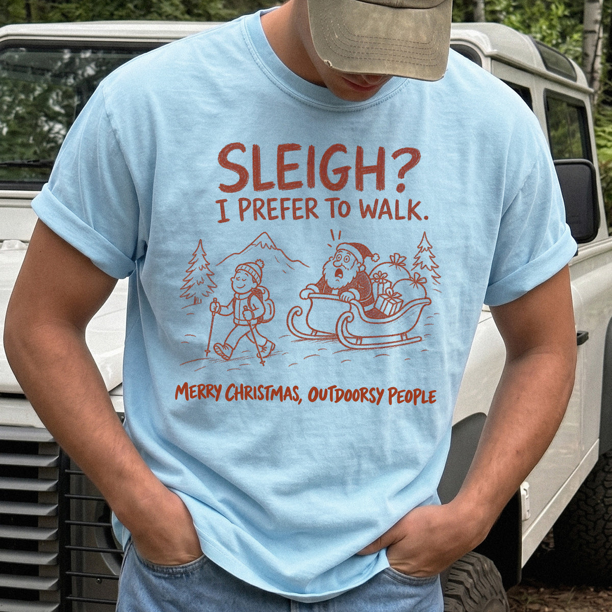 Man wearing a Chambray T-shirt printed with a hiker with trekking poles and a surprised Santa in his sleigh against a mountain backdrop, doodle line-art style
