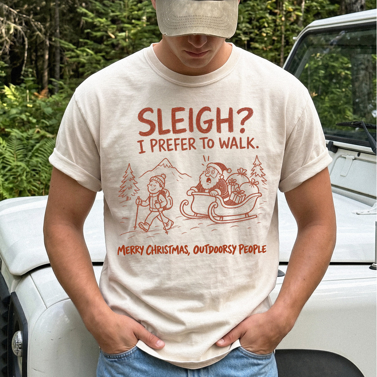 Man wearing an Ivory T-shirt printed with a hiker with trekking poles and a surprised Santa in his sleigh against a mountain backdrop, doodle line-art style