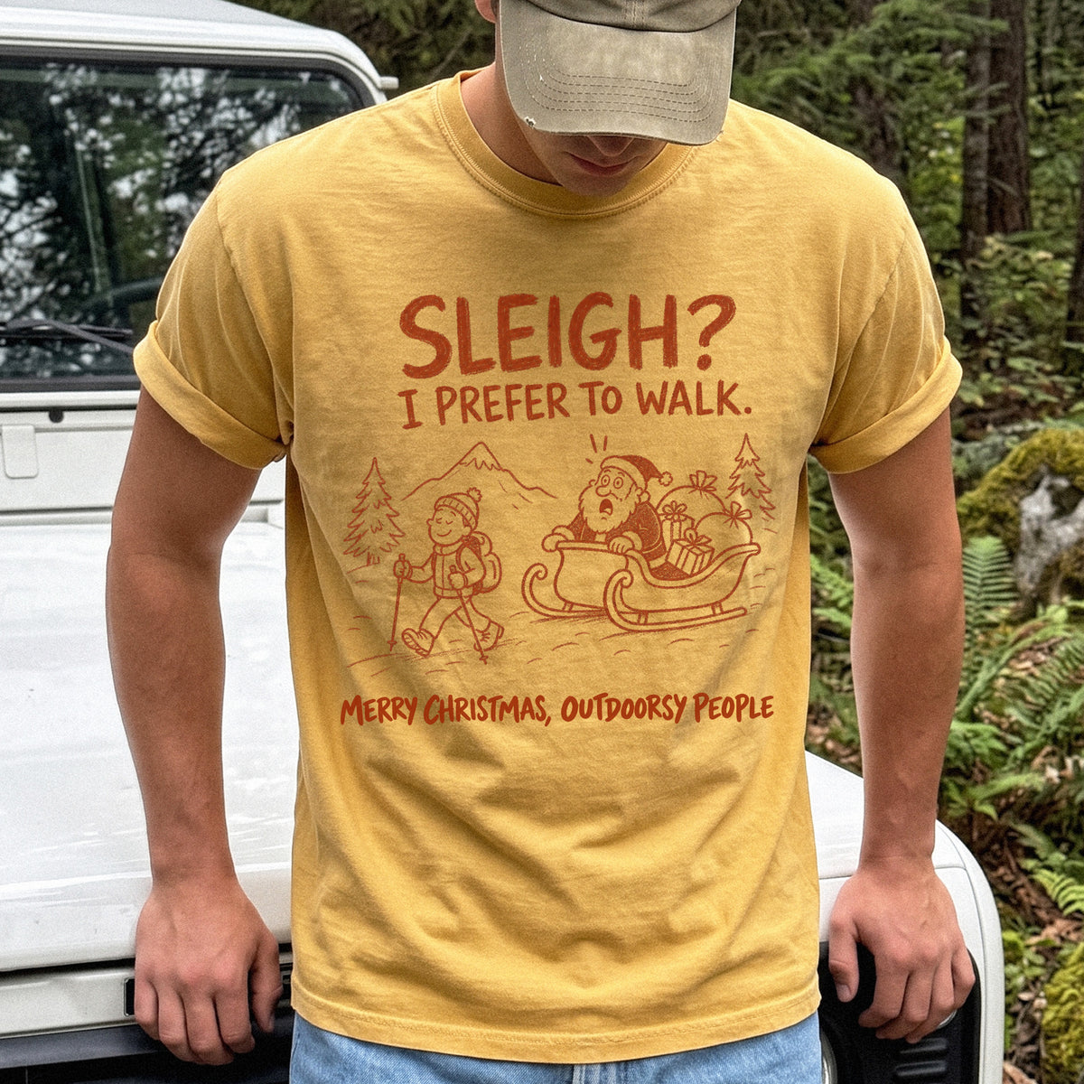 Man wearing a Mustard T-shirt printed with a hiker with trekking poles and a surprised Santa in his sleigh against a mountain backdrop, doodle line-art style