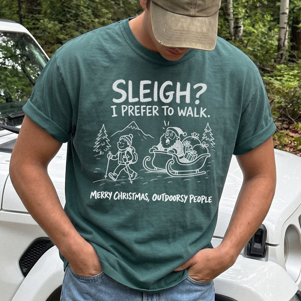 Man wearing a Blue Spruce T-shirt printed with a hiker with trekking poles and a surprised Santa in his sleigh against a mountain backdrop, doodle line-art style