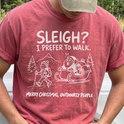 Man wearing a Crimson T-shirt printed with a hiker with trekking poles and a surprised Santa in his sleigh against a mountain backdrop, doodle line-art style