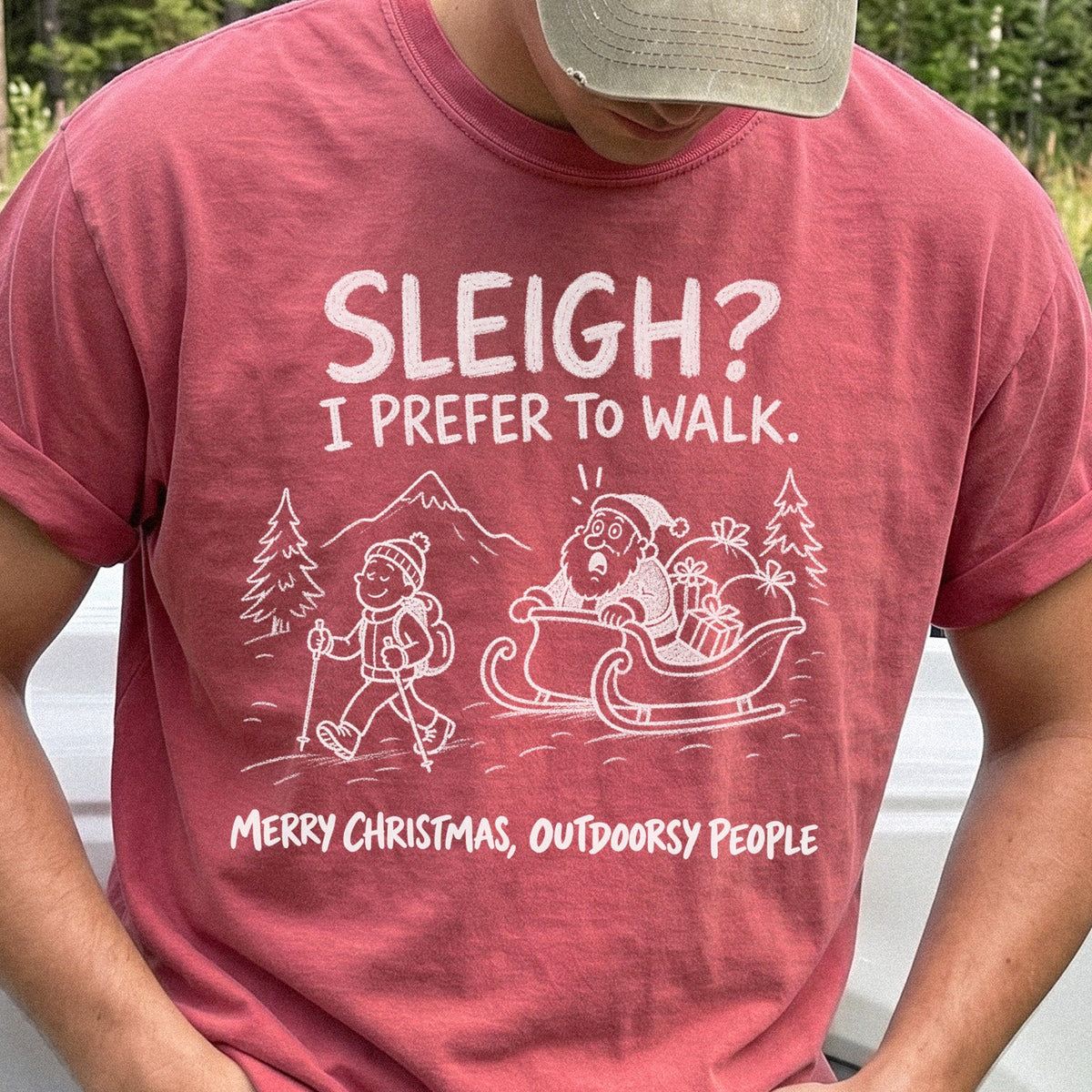 Man wearing a Crimson T-shirt printed with a hiker with trekking poles and a surprised Santa in his sleigh against a mountain backdrop, doodle line-art style