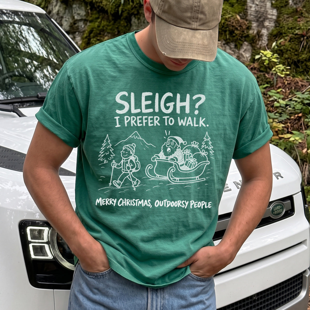 Man wearing a Light Green T-shirt printed with a hiker with trekking poles and a surprised Santa in his sleigh against a mountain backdrop, doodle line-art style