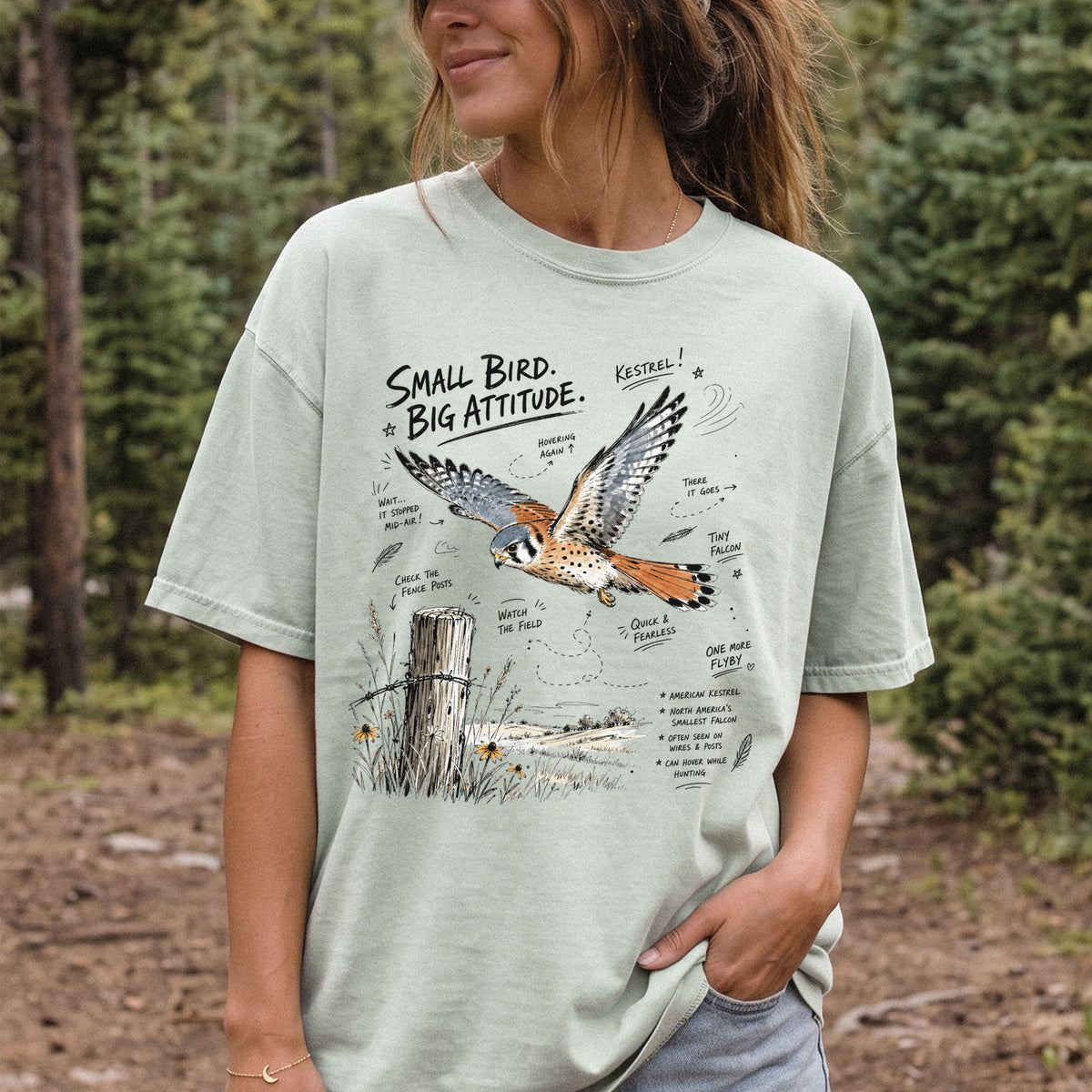 Small Bird, Big Attitude Comfort Colors Shirt – American Kestrel design of an American Kestrel illustrated in mid-flight above an open field, with fence-post details and field-note style callouts, printed on a Bay T-shirt, worn by a woman.