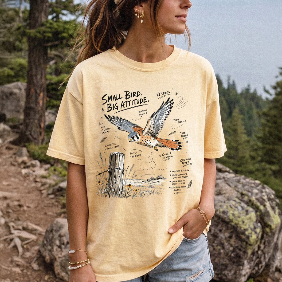 Small Bird, Big Attitude Comfort Colors Shirt – American Kestrel design of an American Kestrel illustrated in mid-flight above an open field, with fence-post details and field-note style callouts, printed on a Butter T-shirt, worn by a woman.