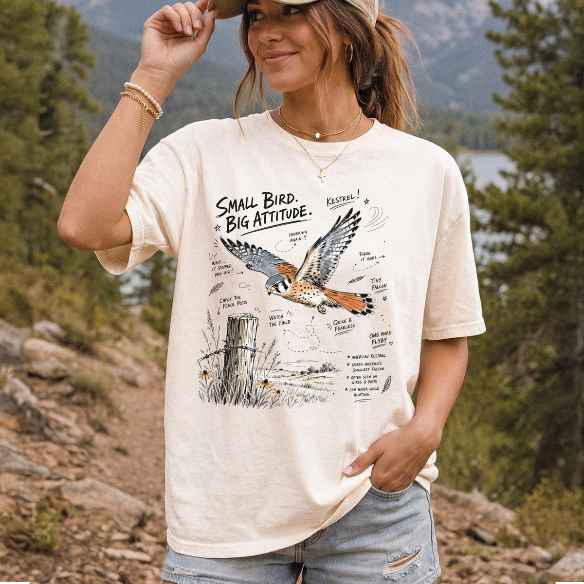 Small Bird, Big Attitude Comfort Colors Shirt – American Kestrel design of an American Kestrel illustrated in mid-flight above an open field, with fence-post details and field-note style callouts, printed on an Ivory T-shirt, worn by a woman.
