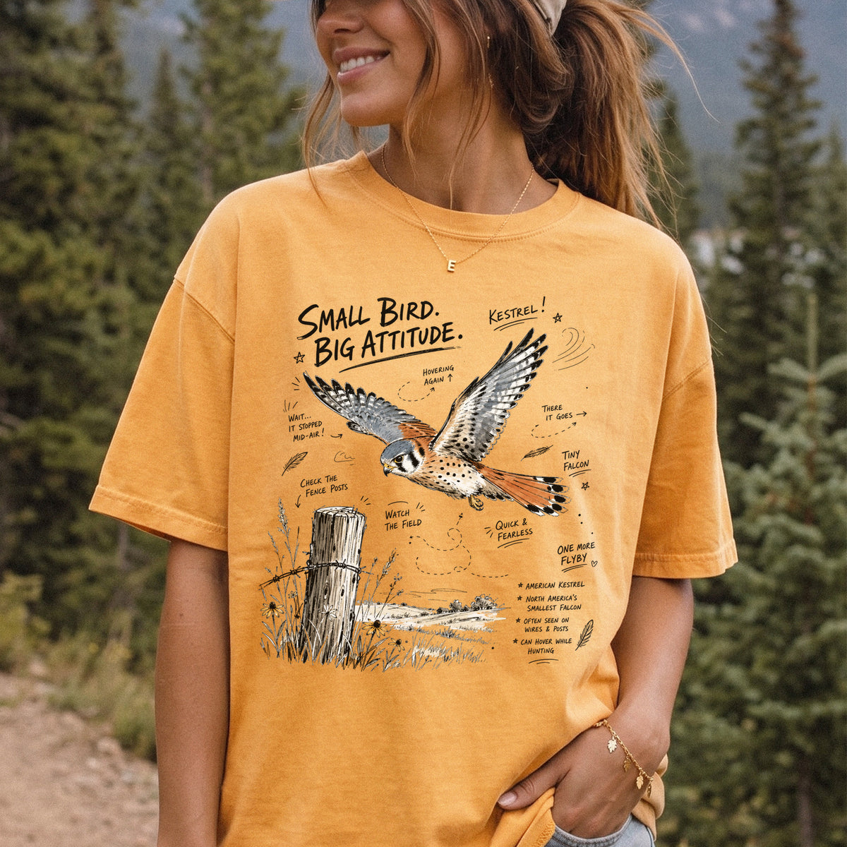 Small Bird, Big Attitude Comfort Colors Shirt – American Kestrel design of an American Kestrel illustrated in mid-flight above an open field, with fence-post details and field-note style callouts, printed on a Mustard T-shirt, worn by a woman.