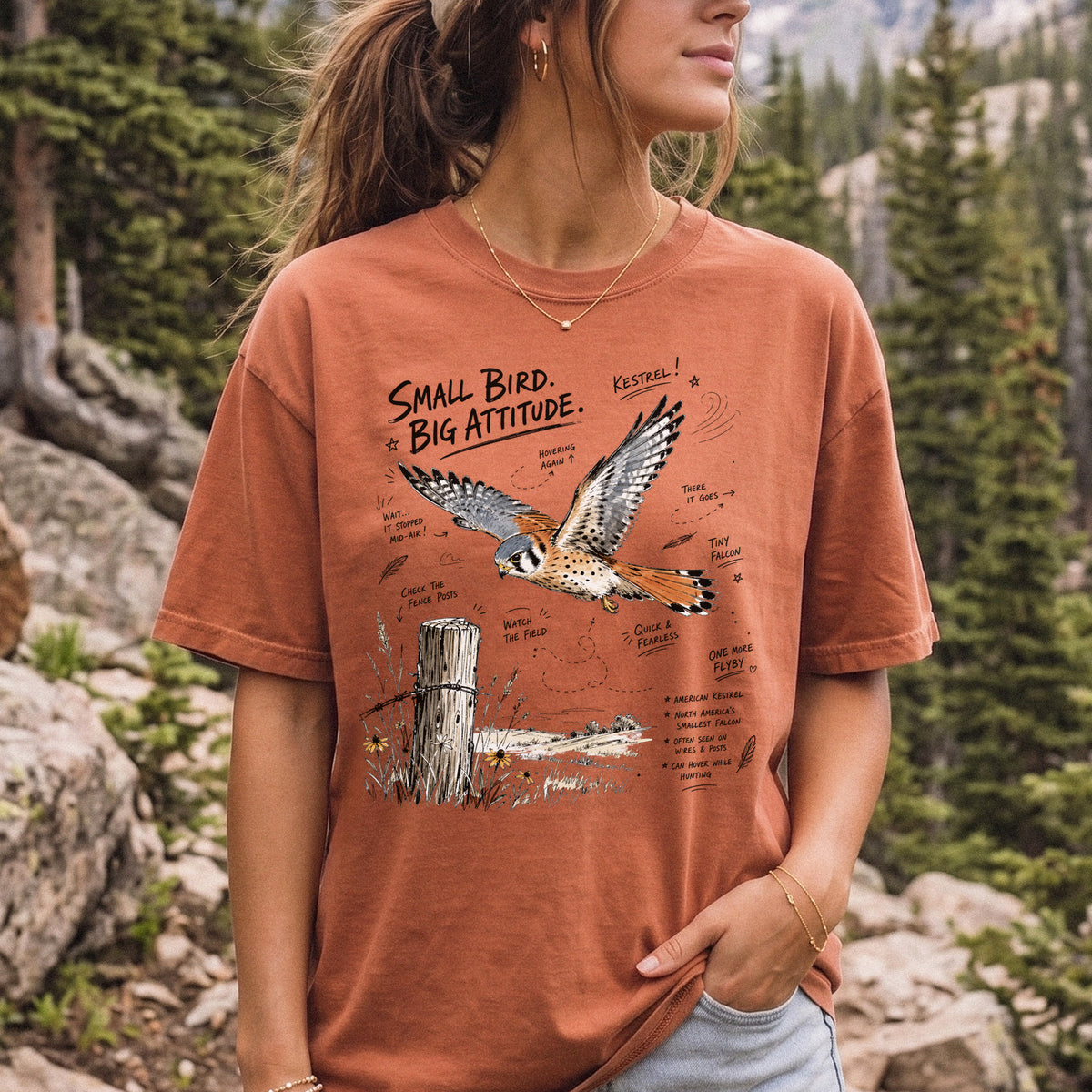 Small Bird, Big Attitude Comfort Colors Shirt – American Kestrel design of an American Kestrel illustrated in mid-flight above an open field, with fence-post details and field-note style callouts, printed on a Yam T-shirt, worn by a woman.