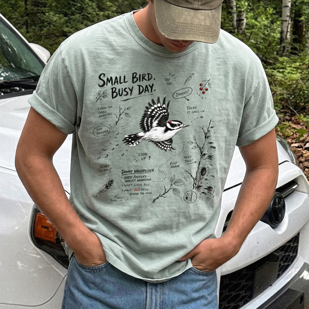Small Bird Busy Day Comfort Colors Shirt – Downy Woodpecker design of a Downy Woodpecker illustrated in motion, along with sketch-style notes, branches, and tree-trunk details, printed on a Bay T-shirt.