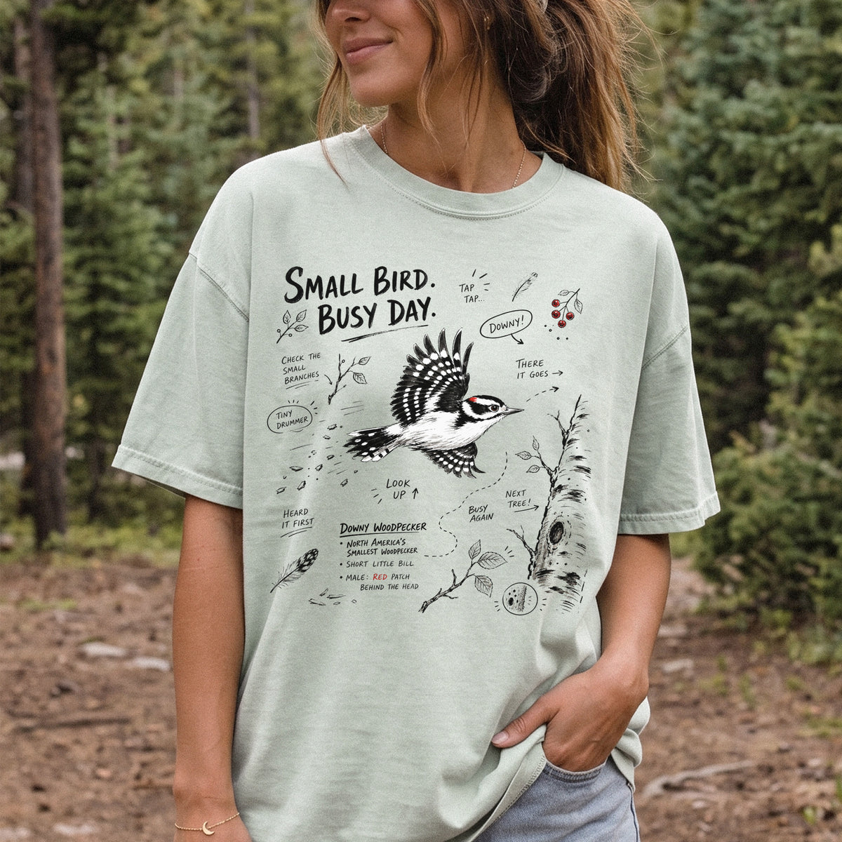 Small Bird Busy Day Comfort Colors Shirt – Downy Woodpecker design of a Downy Woodpecker illustrated in motion, along with sketch-style notes, branches, and tree-trunk details, printed on a Bay T-shirt, worn by a woman.