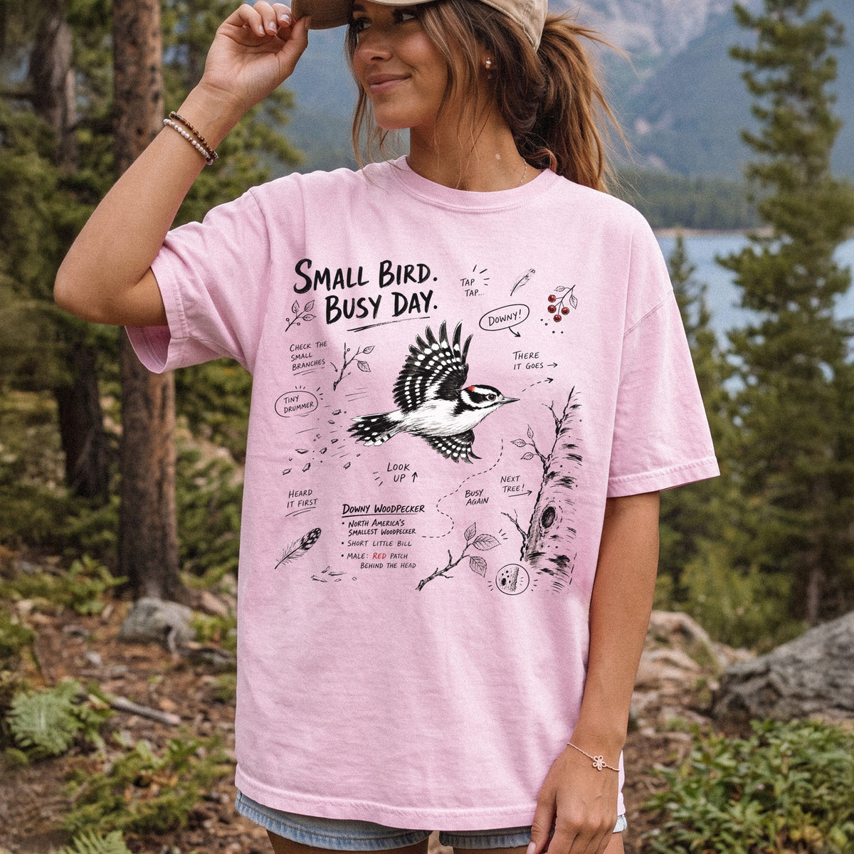 Small Bird Busy Day Comfort Colors Shirt – Downy Woodpecker design of a Downy Woodpecker illustrated in motion, along with sketch-style notes, branches, and tree-trunk details, printed on a Blossom T-shirt, worn by a woman.