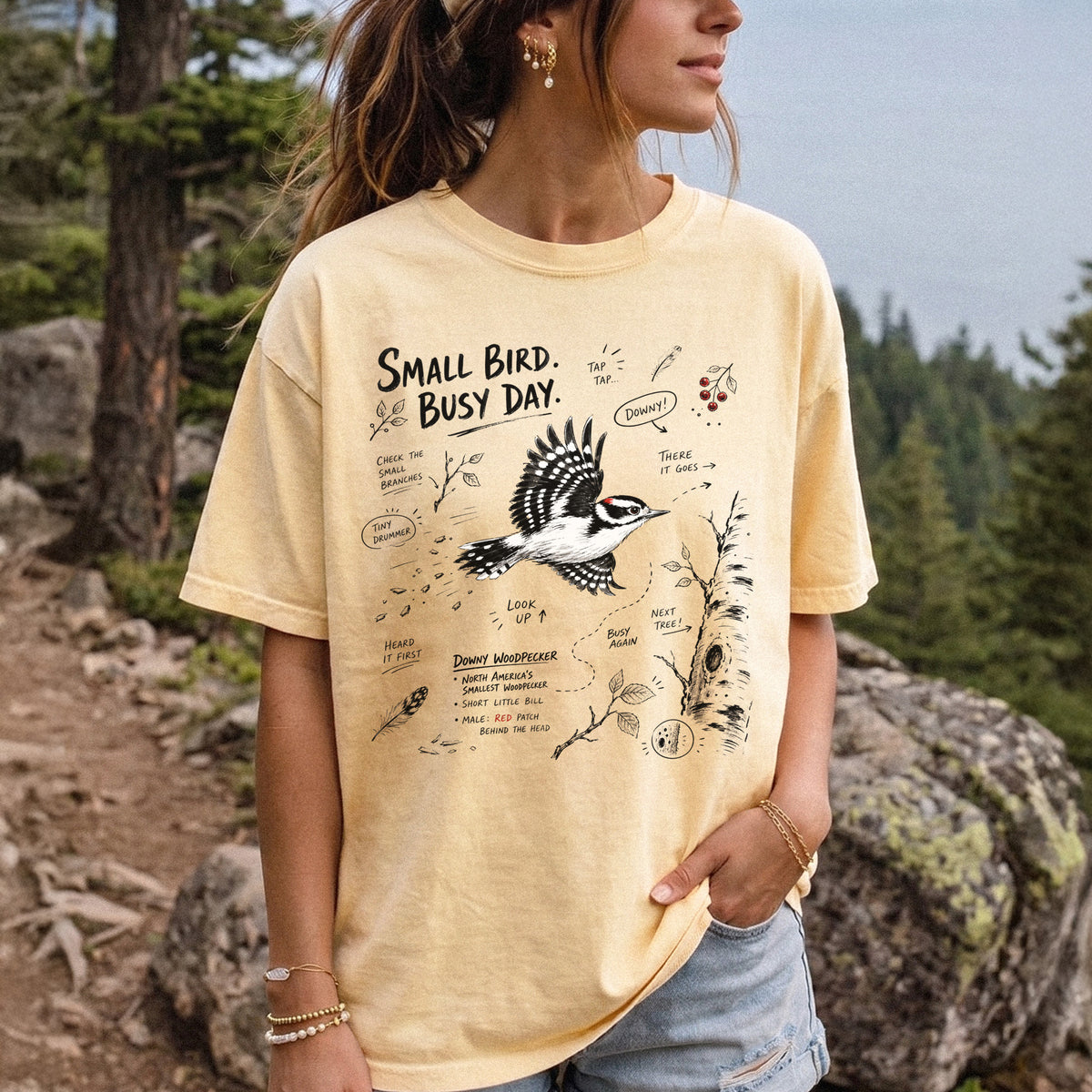 Small Bird Busy Day Comfort Colors Shirt – Downy Woodpecker design of a Downy Woodpecker illustrated in motion, along with sketch-style notes, branches, and tree-trunk details, printed on a Butter T-shirt, worn by a woman.