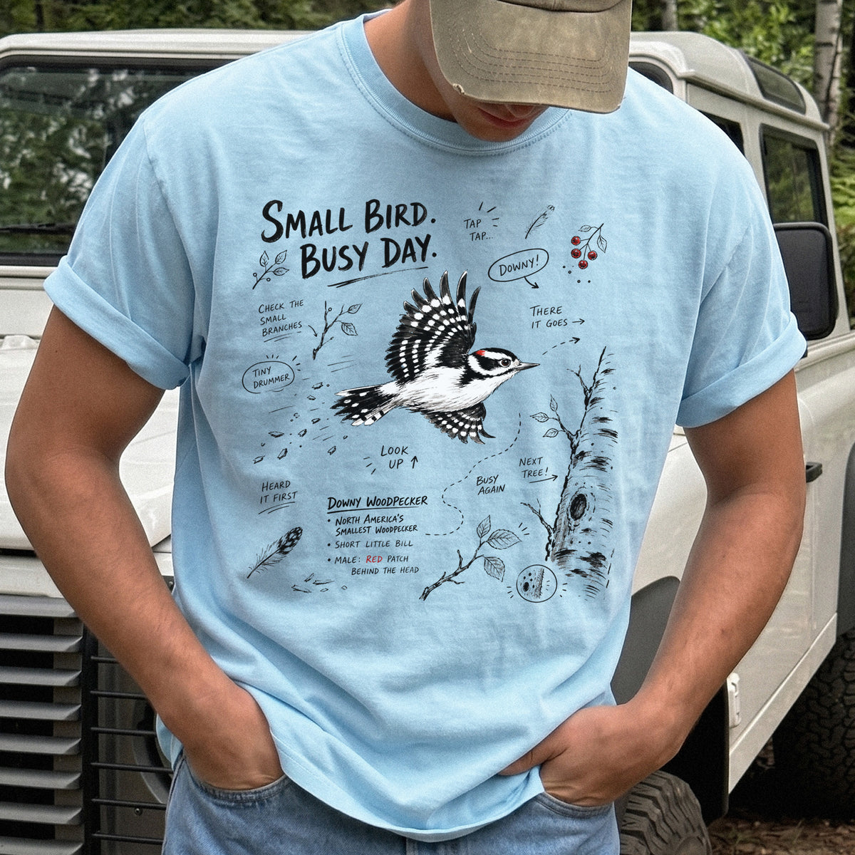 Small Bird Busy Day Comfort Colors Shirt – Downy Woodpecker design of a Downy Woodpecker illustrated in motion, along with sketch-style notes, branches, and tree-trunk details, printed on a Chambray T-shirt.