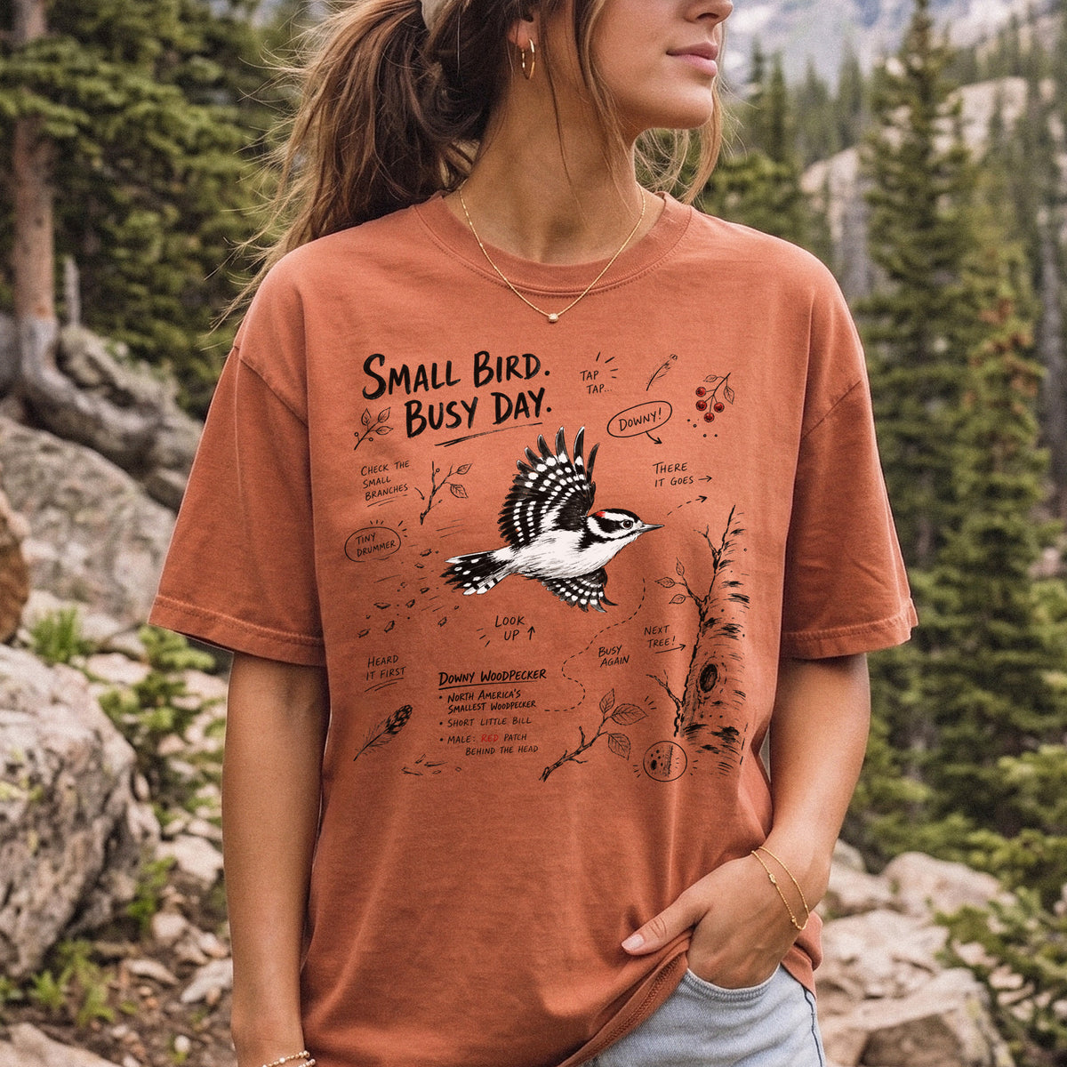 Small Bird Busy Day Comfort Colors Shirt – Downy Woodpecker design of a Downy Woodpecker illustrated in motion, along with sketch-style notes, branches, and tree-trunk details, printed on a Yam T-shirt, worn by a woman.