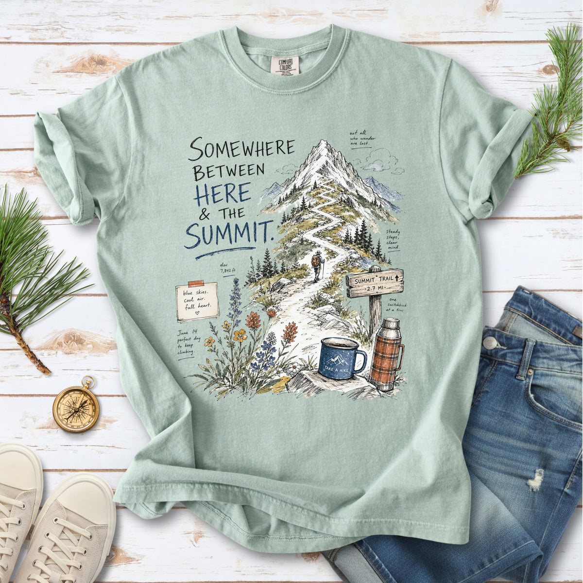A hand-drawn mountain summit trail scene with wildflowers, a trail sign, a hiker, and a camp mug design on a Bay T-shirt