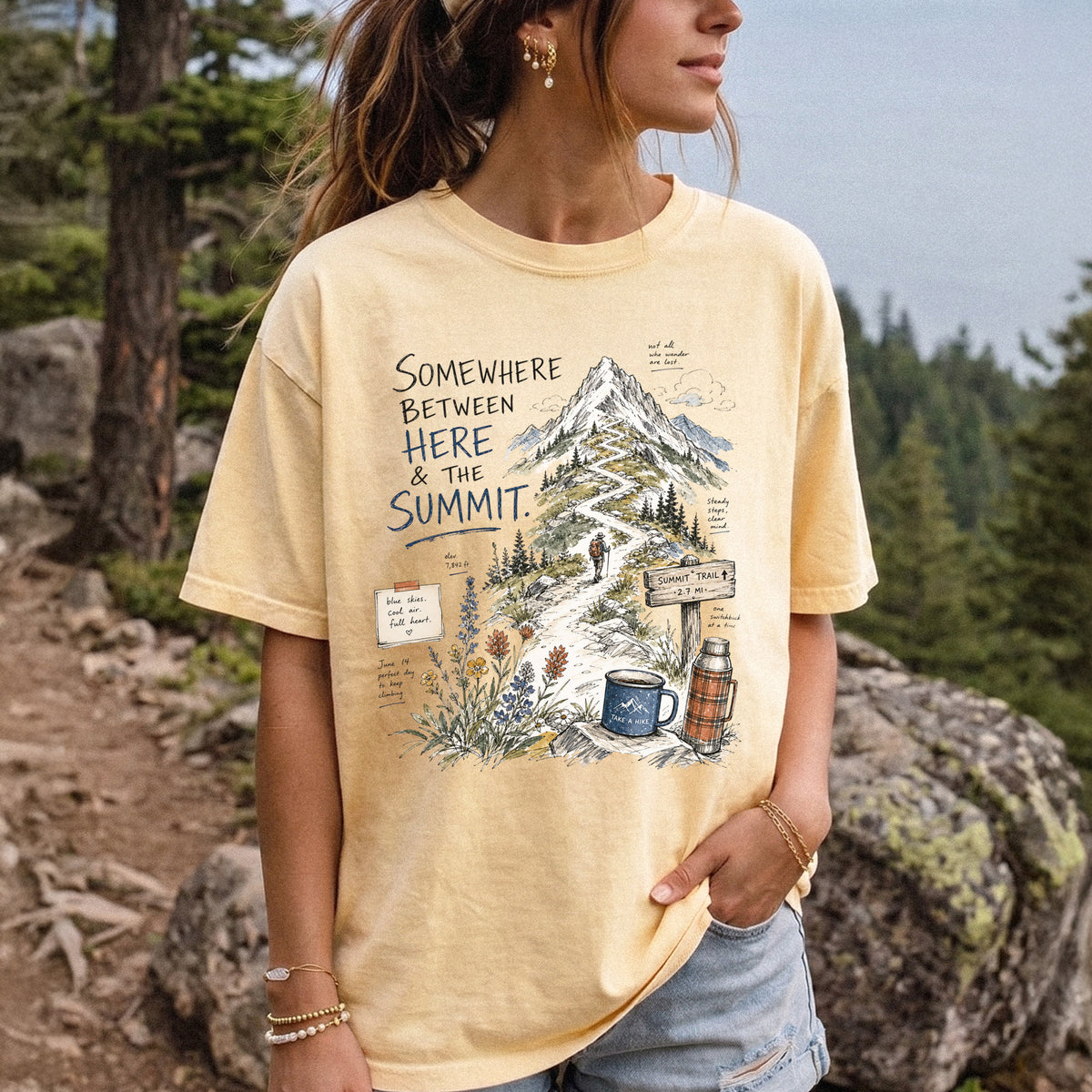 Woman wearing a Butter T-shirt printed with a hand-drawn mountain summit trail scene with wildflowers, a trail sign, a hiker, and a camp mug